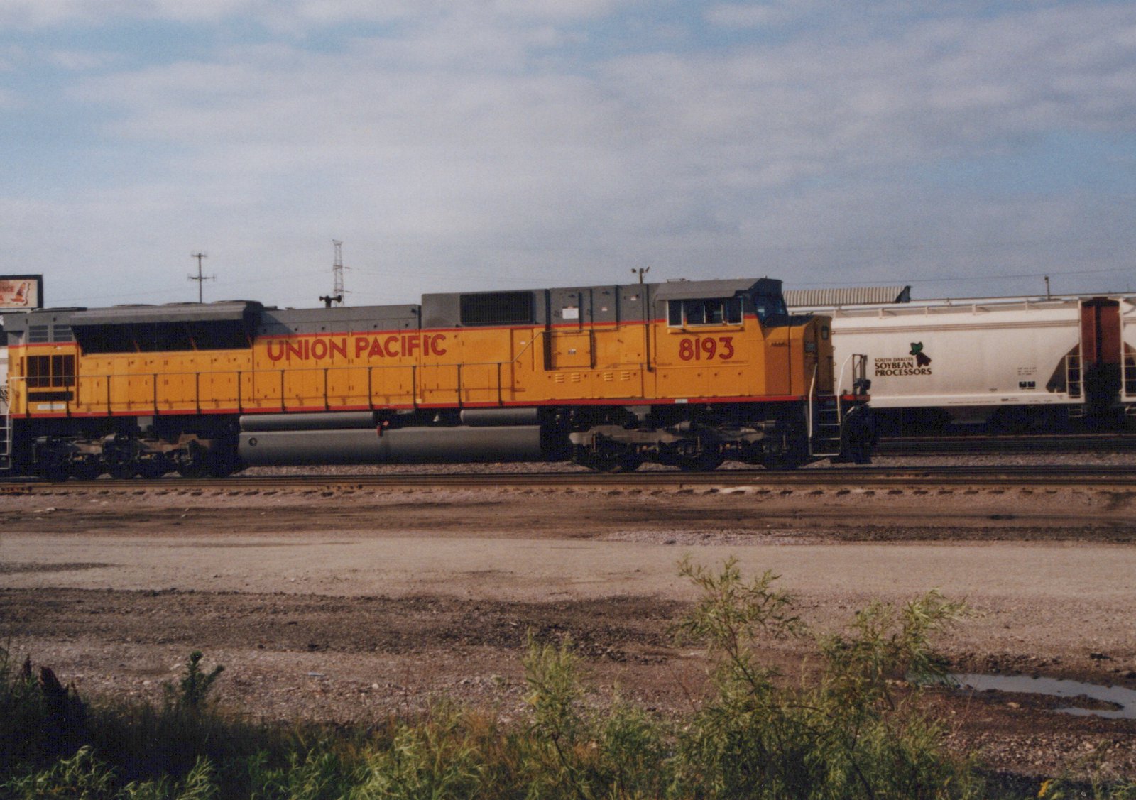 A yellow Union Pacific locomotive numbered 8193 is coupled to a white freight car with 'South Dakota Soybean Processors' lettering, on a set of railroad tracks in a gravel yard under an overcast sky.