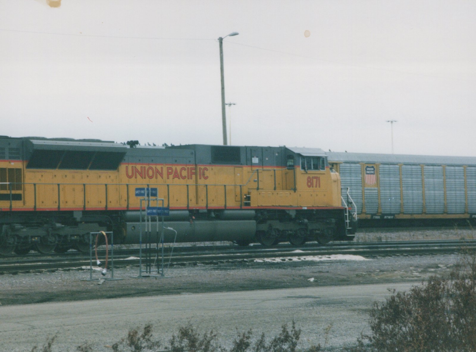 A yellow and gray Union Pacific locomotive numbered 8171 is parked on the tracks next to a freight car in a railyard with snow on the ground.