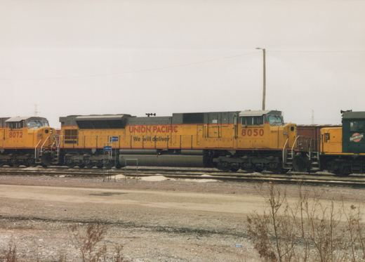 A side view of two yellow Union Pacific locomotives, numbered 8050 and 8072, with a green Chicago and North Western caboose in the background, parked on tracks at Proviso Yard on an overcast winter day.