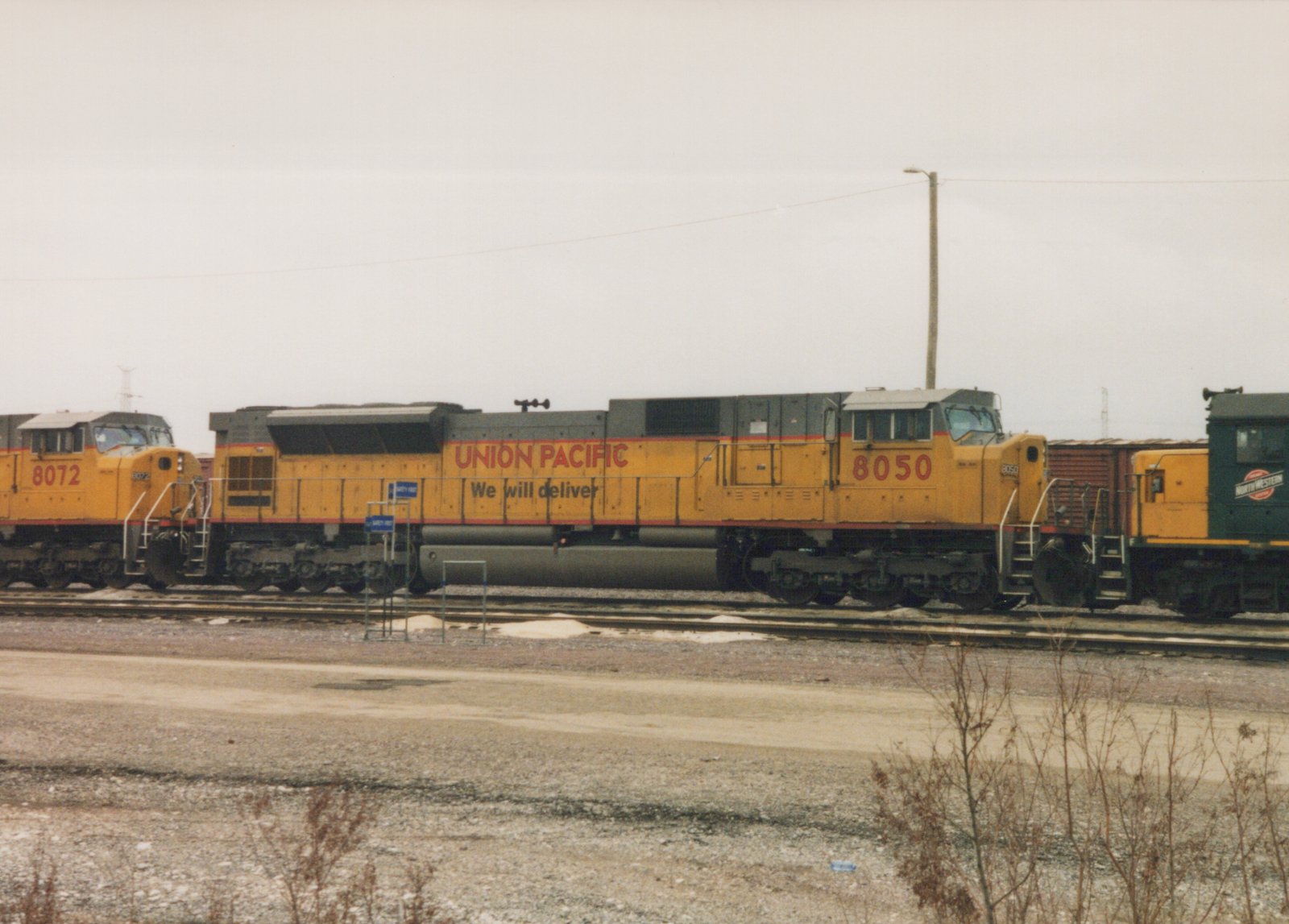 A side view of two yellow Union Pacific locomotives, numbered 8050 and 8072, with a green Chicago and North Western caboose in the background, parked on tracks at Proviso Yard on an overcast winter day.