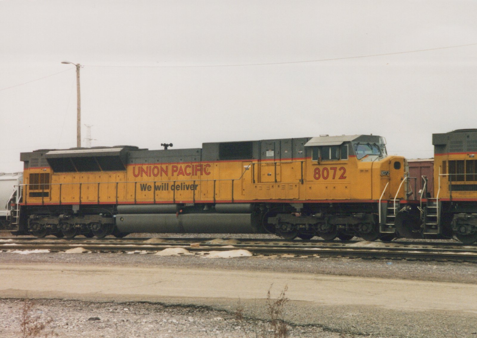 A yellow Union Pacific locomotive numbered 8072 is parked on tracks in a rail yard, with snow on the ground and an overcast sky.