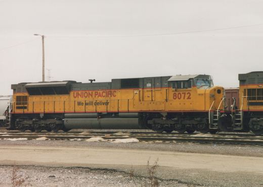 A yellow Union Pacific locomotive numbered 8072 is parked on tracks in a rail yard, with snow on the ground and an overcast sky.