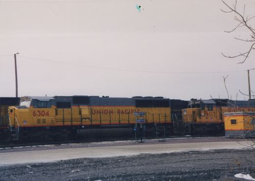 Side view of two yellow and gray Union Pacific diesel locomotives, numbered 6304 and 4117, standing on tracks in a yard with snow on the ground.
