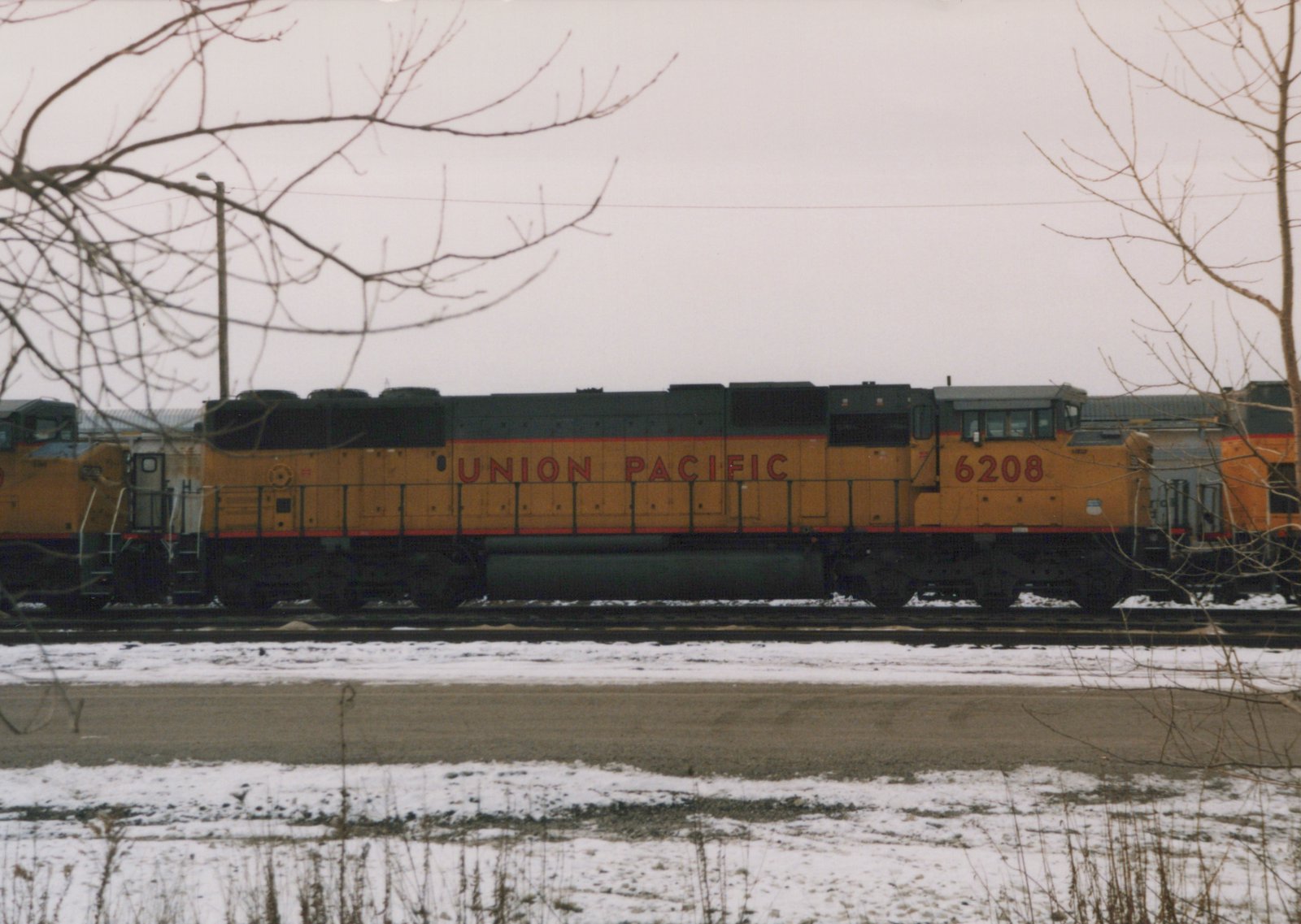 A side view of a Union Pacific diesel locomotive, number 6208, painted yellow with black and red accents, positioned on tracks with snow on the ground and bare trees in the foreground.