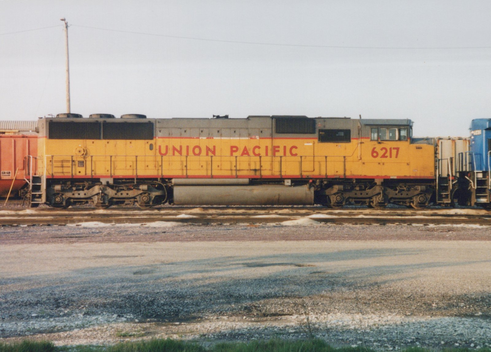 A yellow and gray Union Pacific locomotive numbered 6217 sits on tracks in a gravel yard, with freight cars and another blue locomotive visible in the background under an overcast sky.