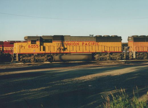 Side view of a yellow Union Pacific diesel locomotive, number 6055, in a rail yard with other locomotives nearby and a clear sky.