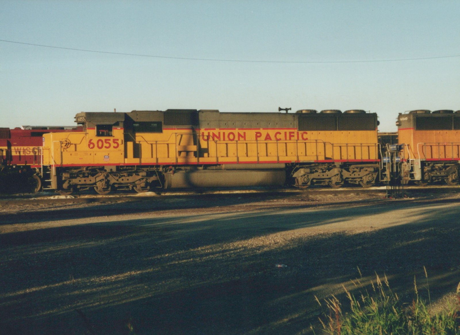 Side view of a yellow Union Pacific diesel locomotive, number 6055, in a rail yard with other locomotives nearby and a clear sky.