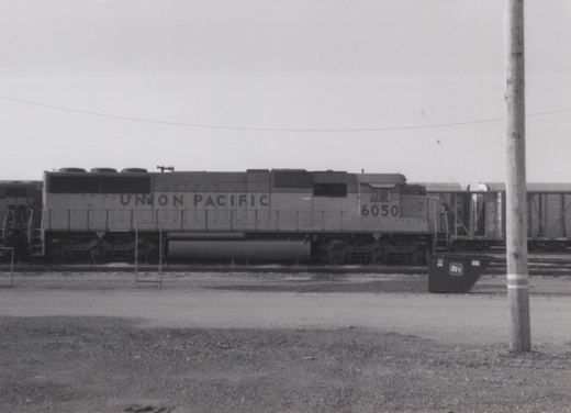A black-and-white photo of a Union Pacific diesel locomotive, number 6050, parked on a track in a rail yard with boxcars behind it and a BFI dumpster in the foreground.