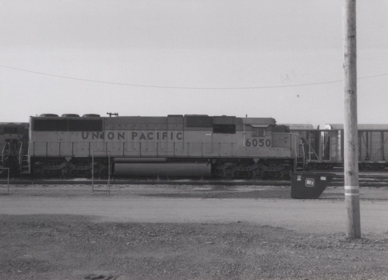 A black-and-white photo of a Union Pacific diesel locomotive, number 6050, parked on a track in a rail yard with boxcars behind it and a BFI dumpster in the foreground.