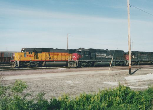 Three diesel locomotives, one in Union Pacific yellow and two in Southern Pacific black livery, are parked in a yard with green bushes in the foreground.
