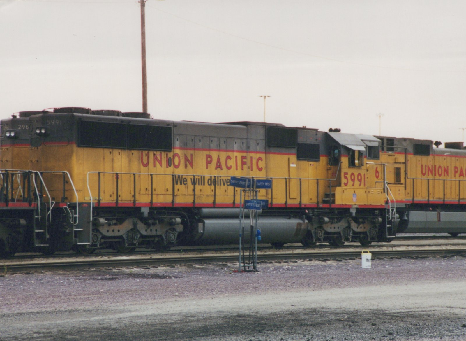 Two yellow and gray Union Pacific locomotives, numbers 5991 and 296, are parked side by side in a gravel yard on a cloudy day.