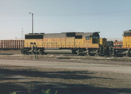 A yellow and gray Union Pacific diesel locomotive, number 5948, sits on railroad tracks in a yard with other freight cars under a clear sky.