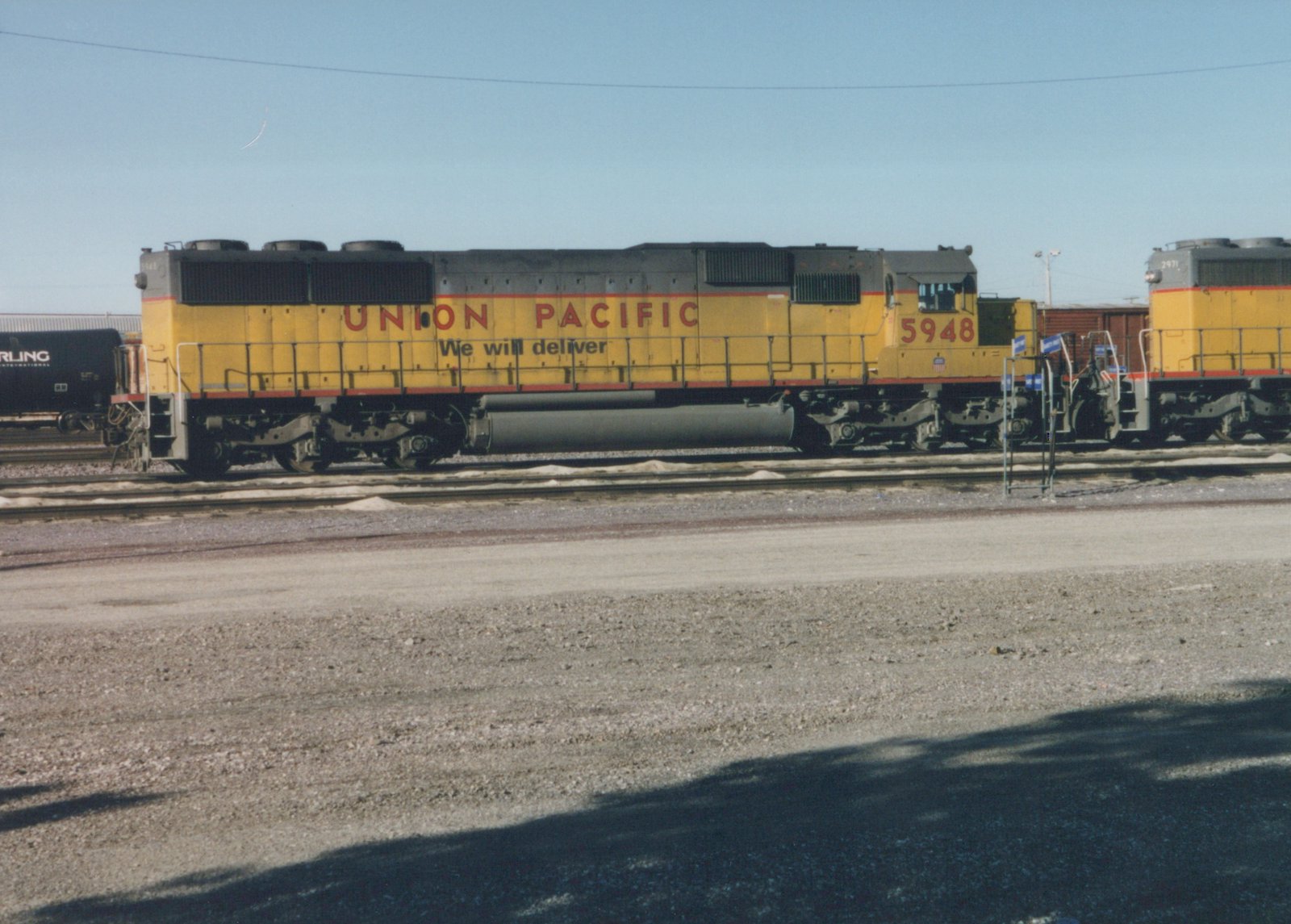 Side view of a yellow Union Pacific diesel locomotive #5948 parked in a railyard with snow on the ground, with another similar locomotive #2971 and a black freight car in the background under a clear sky.