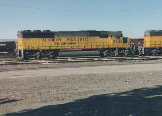 Side view of a yellow Union Pacific diesel locomotive #5948 parked in a railyard with snow on the ground, with another similar locomotive #2971 and a black freight car in the background under a clear sky.