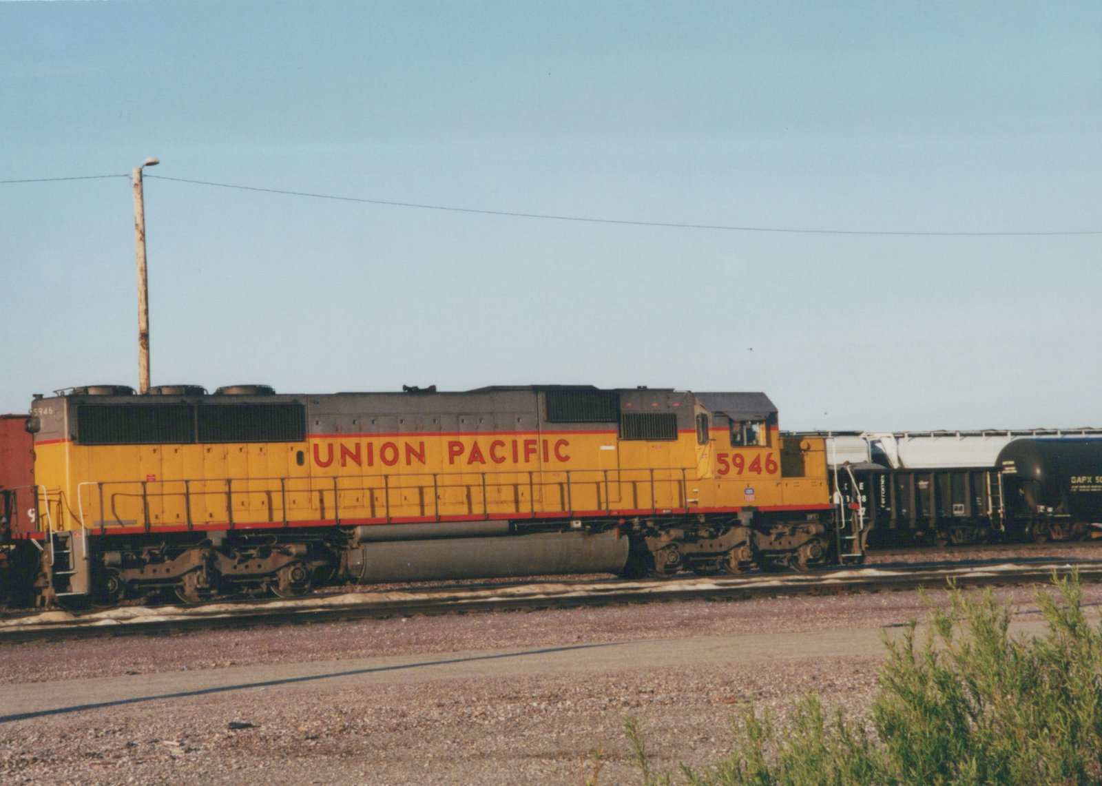 Side view of a yellow and red Union Pacific locomotive numbered 5946, stopped on railroad tracks next to other freight cars.