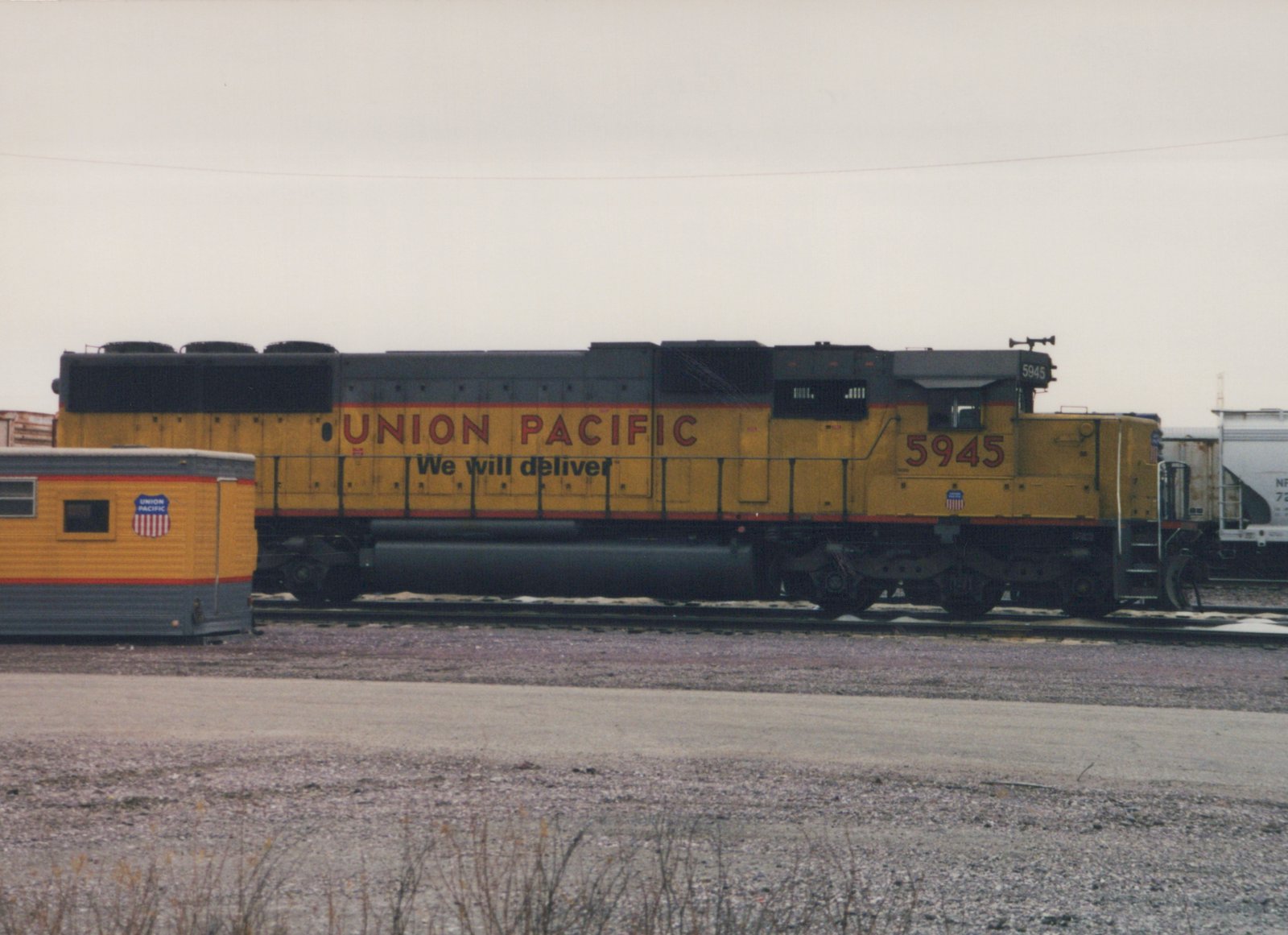 Side view of a yellow Union Pacific locomotive numbered 5945, parked on tracks in a gravel yard with a trailer car nearby.