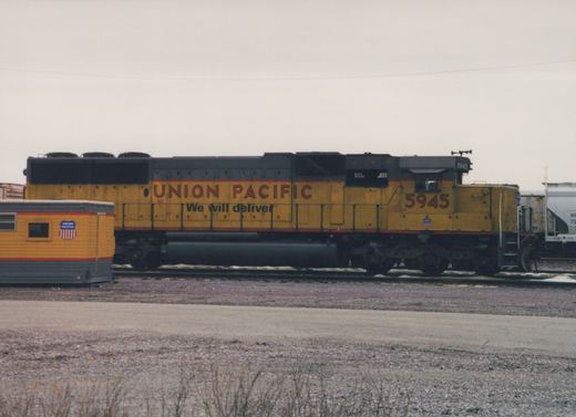 Side view of a yellow Union Pacific locomotive numbered 5945, parked on tracks in a gravel yard with a trailer car nearby.