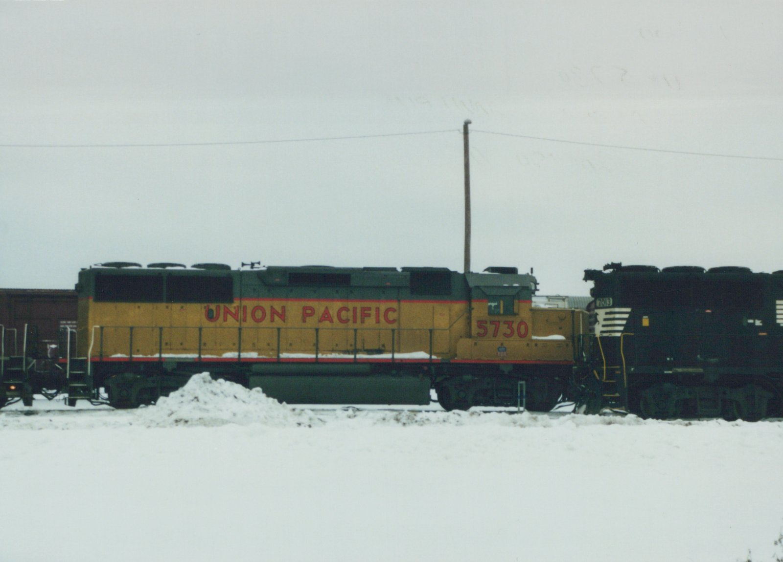 Side-view photograph of two Union Pacific diesel locomotives, numbers 5730 and 7013, on snow-covered tracks in a yard under an overcast sky.