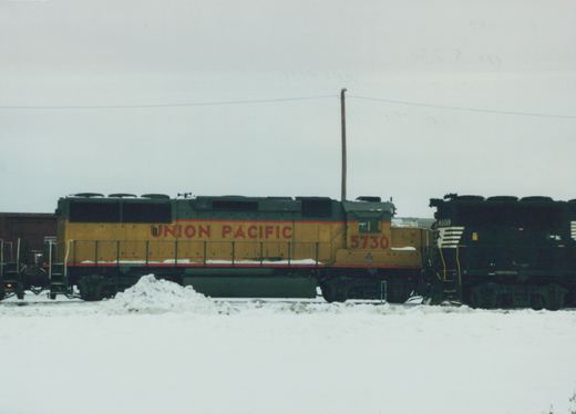 Side-view photograph of two Union Pacific diesel locomotives, numbers 5730 and 7013, on snow-covered tracks in a yard under an overcast sky.