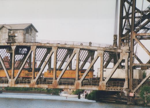 A yellow Union Pacific diesel locomotive crosses a steel truss bridge over a river, with boats moored below and a small building on the bridge's structure.