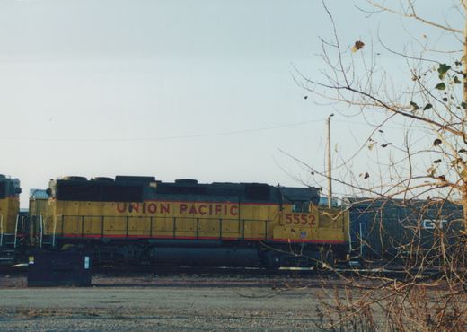 A yellow Union Pacific locomotive with the number 5552 on its side is parked on tracks in a yard, with a bare tree in the foreground.