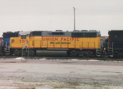 Side view of a yellow and gray Union Pacific diesel locomotive number 5517 standing on tracks in a gravel yard, with two workers in hard hats nearby and other freight cars in the background.