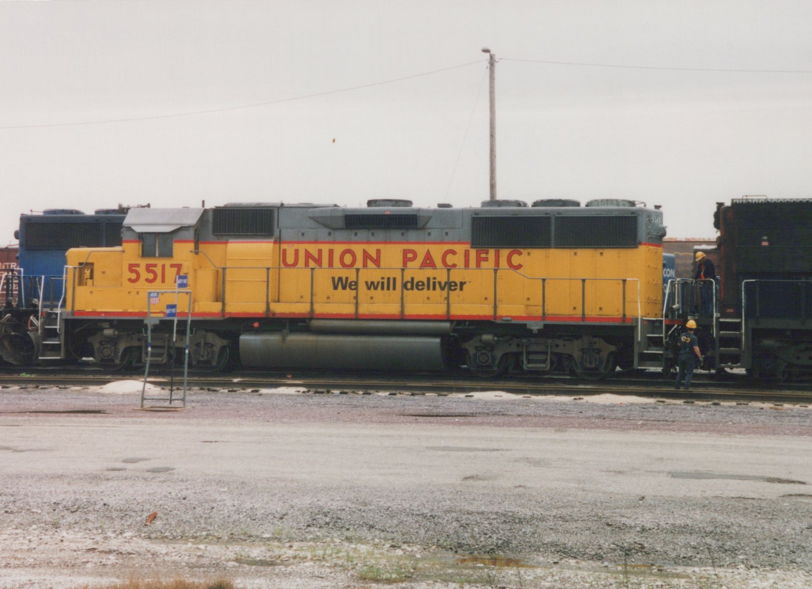 Side view of a yellow and gray Union Pacific diesel locomotive number 5517 standing on tracks in a gravel yard, with two workers in hard hats nearby and other freight cars in the background.