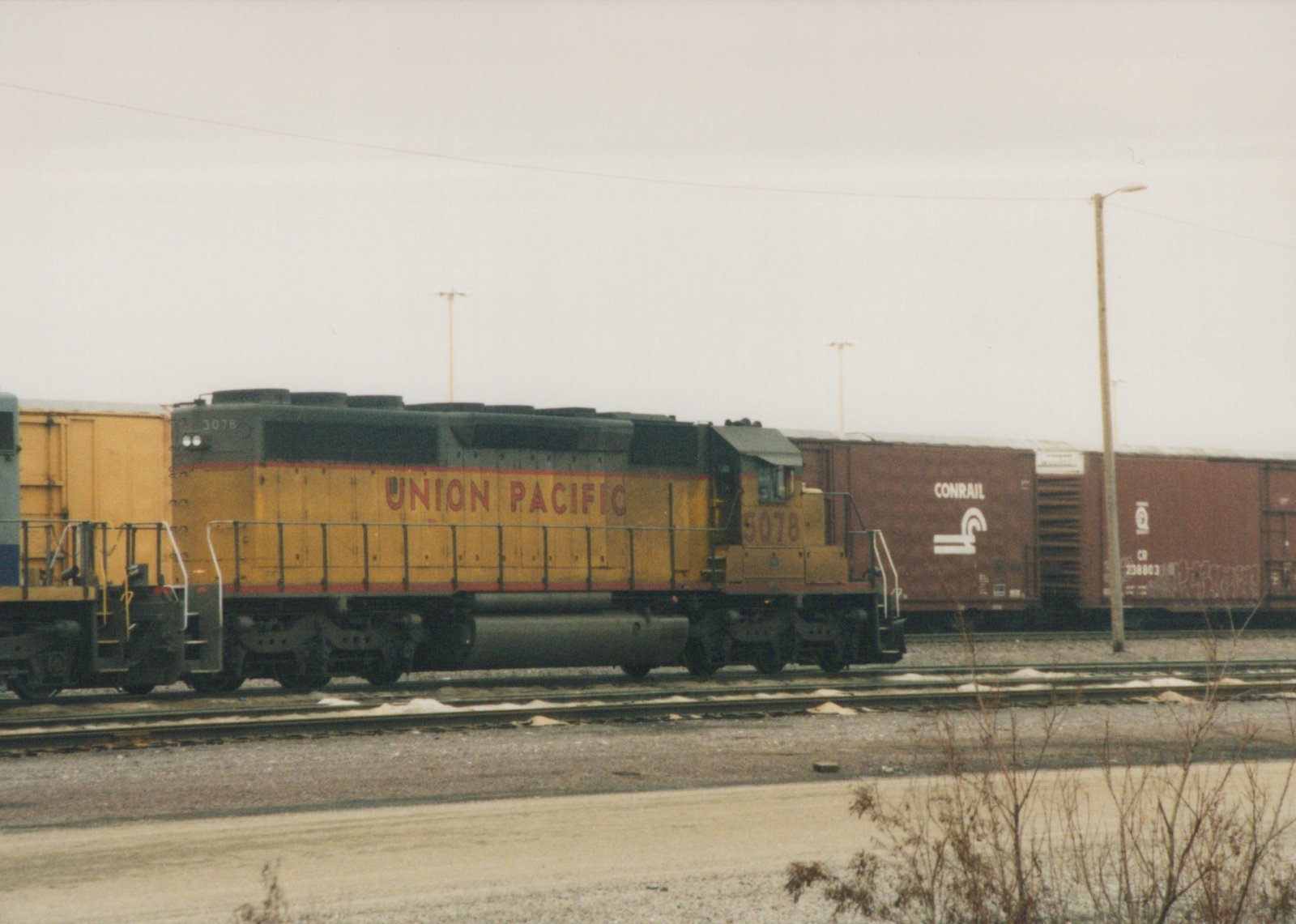 A yellow Union Pacific diesel locomotive with the number 5078 is parked on railroad tracks next to brown Conrail freight cars in a yard on a cloudy winter day.