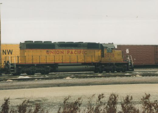 Side view of a yellow and red Union Pacific diesel locomotive, number 3078, on tracks in a rail yard with boxcars behind it, under an overcast sky.