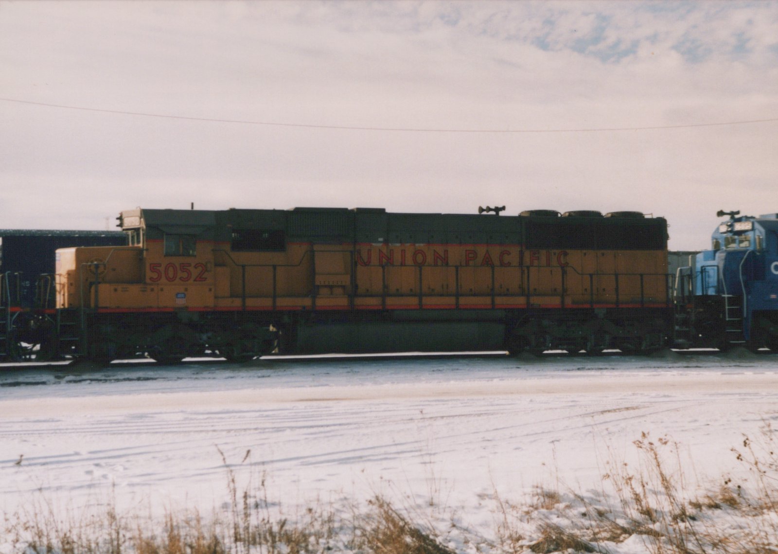 A yellow and black Union Pacific diesel locomotive numbered 5052 stands on snowy tracks, with a blue Conrail locomotive beside it in a rail yard under an overcast sky.