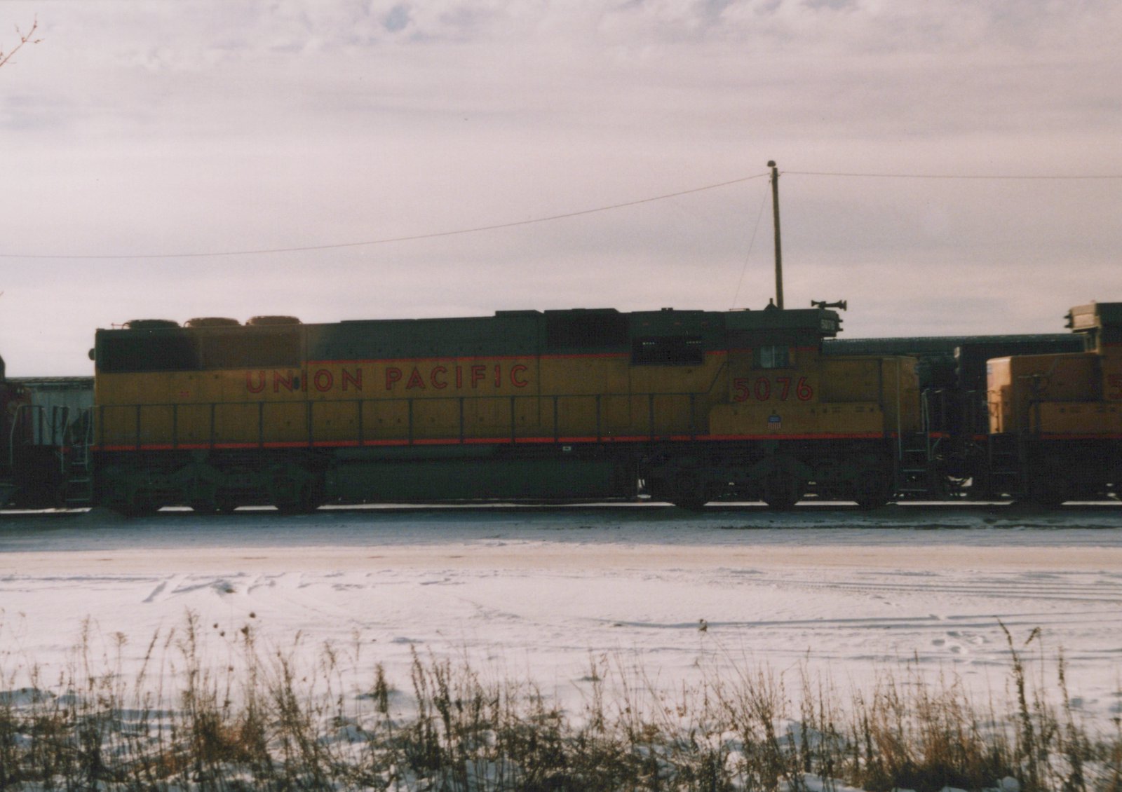 A yellow and green Union Pacific diesel locomotive numbered 5076 is parked on a snow-covered railroad yard under an overcast sky.