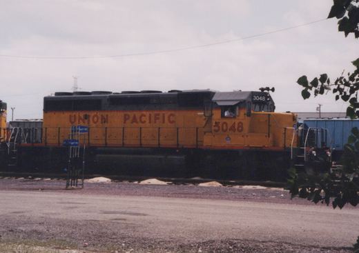 A yellow and black Union Pacific diesel locomotive numbered 3048 is parked on a railroad track next to a gravel lot, with a blue freight car and some trees nearby.