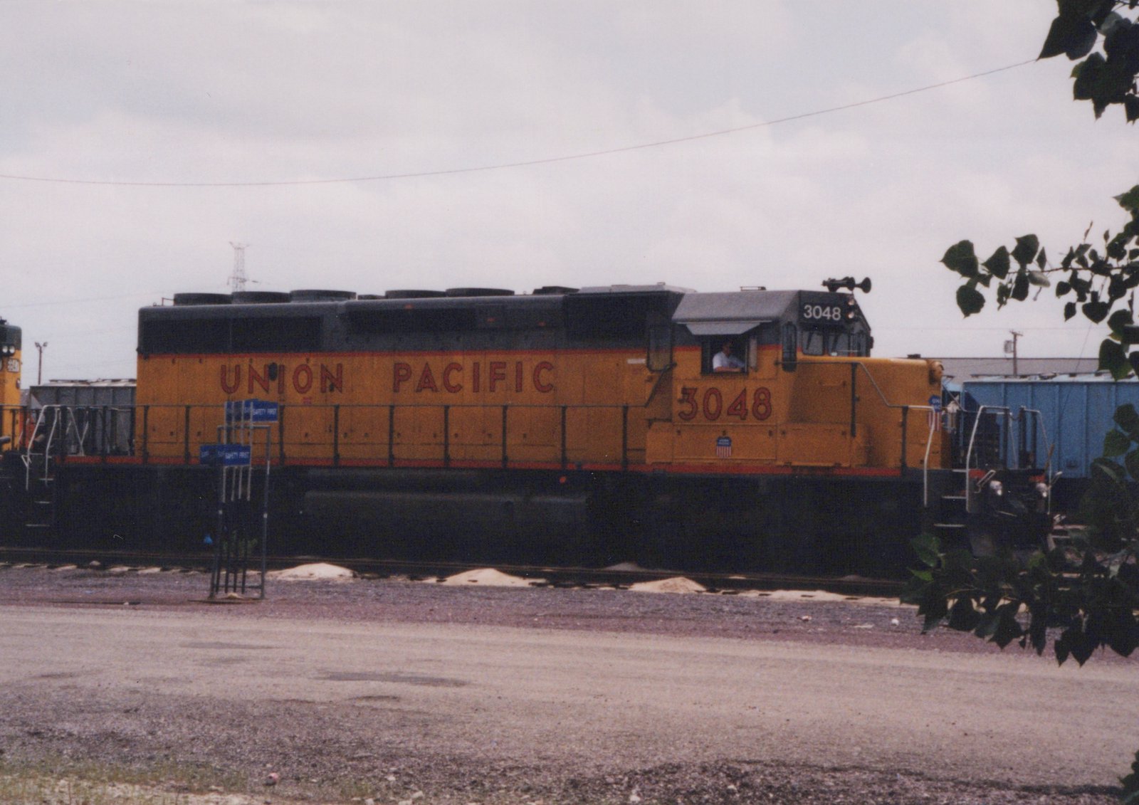 A yellow and black Union Pacific diesel locomotive numbered 3048 is parked on a railroad track next to a gravel lot, with a blue freight car and some trees nearby.