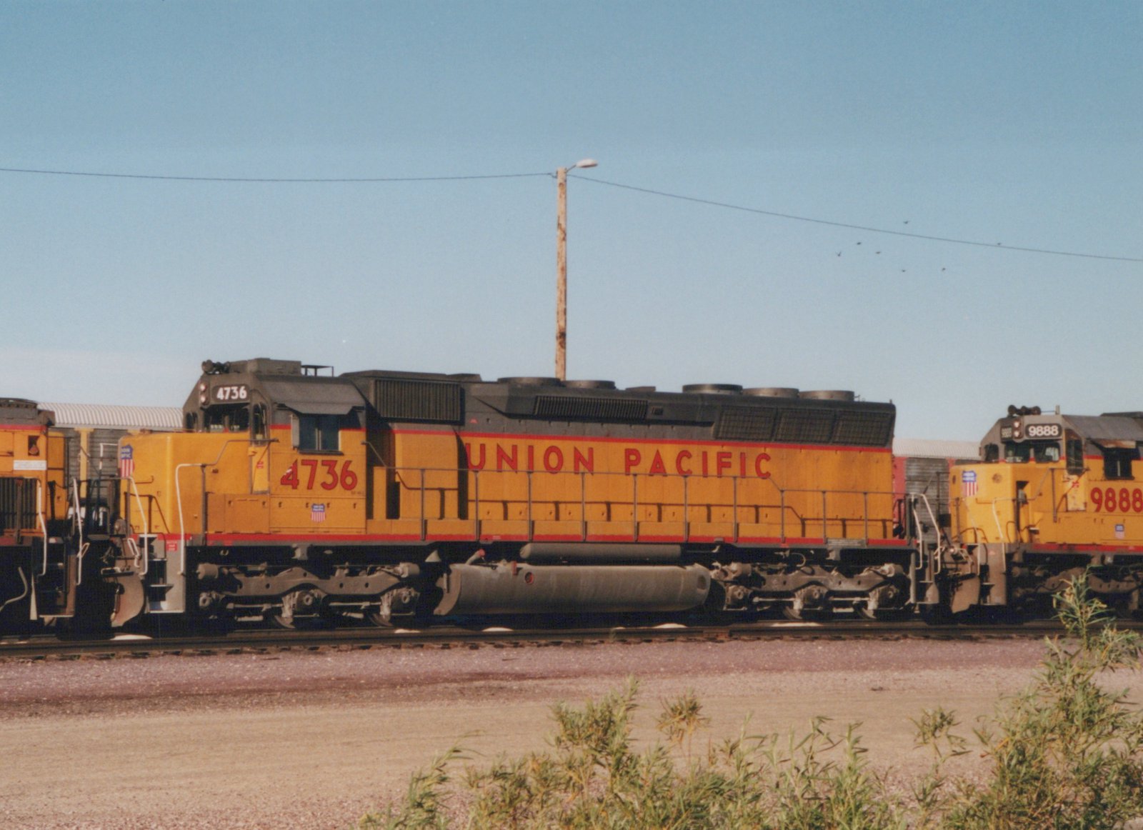 Two yellow Union Pacific locomotives, numbers 4736 and 9888, are parked side by side on railroad tracks in a gravel yard under a clear blue sky.