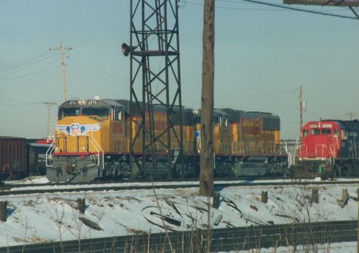 A winter scene at a rail yard showing three diesel locomotives: a yellow SD40-2 numbered 4576 leads another yellow SD40-2 and a red GP38-2, all in Union Pacific livery, with snow on the ground and freight cars nearby.
