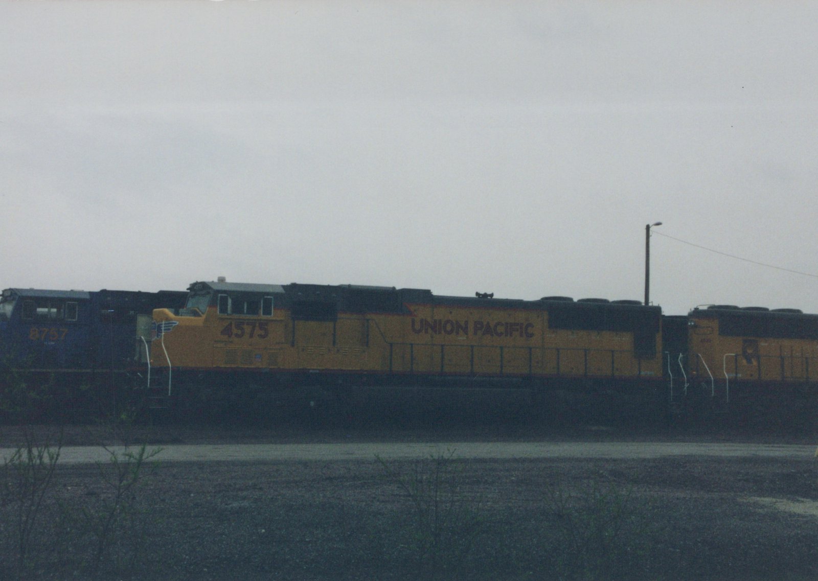 Side view of a yellow-and-gray Union Pacific diesel locomotive numbered 4575, coupled to a second yellow unit, with a dark blue locomotive numbered 8757 at left, under an overcast sky with gravel and bare shrubs in front.