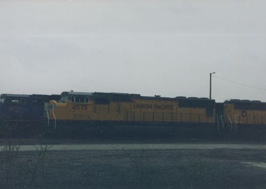 Side view of a yellow-and-gray Union Pacific diesel locomotive numbered 4575, coupled to a second yellow unit, with a dark blue locomotive numbered 8757 at left, under an overcast sky with gravel and bare shrubs in front.