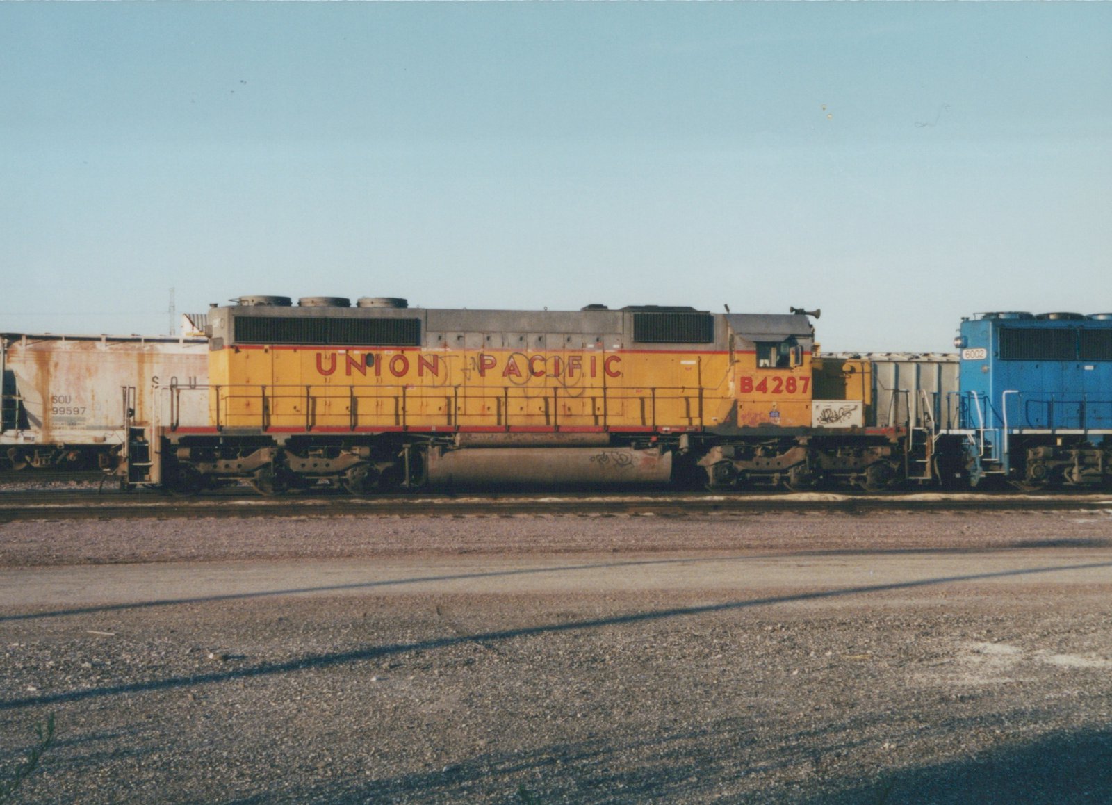 Side view of a weathered yellow Union Pacific diesel locomotive numbered B4287 on a yard track, with a rusty covered hopper on the left and the front of a blue locomotive numbered 6002 on the right, under a clear sky.
