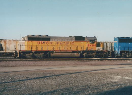 Side view of a weathered yellow Union Pacific diesel locomotive numbered B4287 on a yard track, with a rusty covered hopper on the left and the front of a blue locomotive numbered 6002 on the right, under a clear sky.
