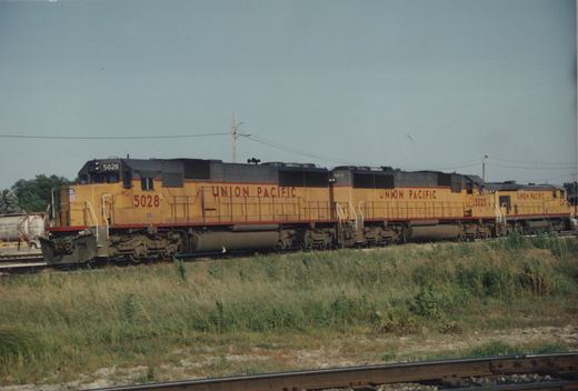 Three yellow-and-gray Union Pacific diesel locomotives stand on a track beyond a grassy embankment, with a container car at the left.