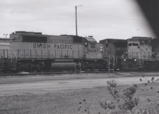 Black-and-white side view of a six-axle Union Pacific diesel locomotive numbered B4240, parked in a rail yard with other locomotives under an overcast sky, with weeds in the foreground.