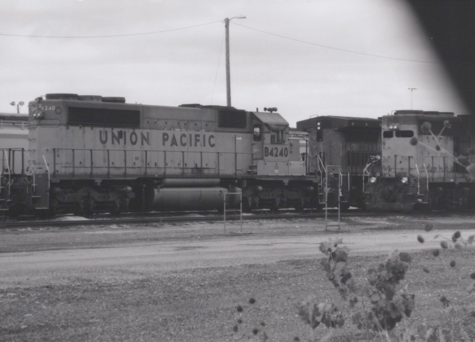 Black-and-white side view of a six-axle Union Pacific diesel locomotive numbered B4240, parked in a rail yard with other locomotives under an overcast sky, with weeds in the foreground.