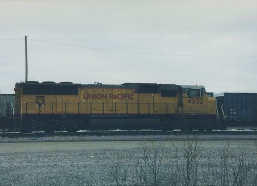 Side view of a yellow Union Pacific diesel locomotive numbered 4232 standing on yard tracks, with a blue hopper car behind it, patches of snow and bare shrubs in the foreground.