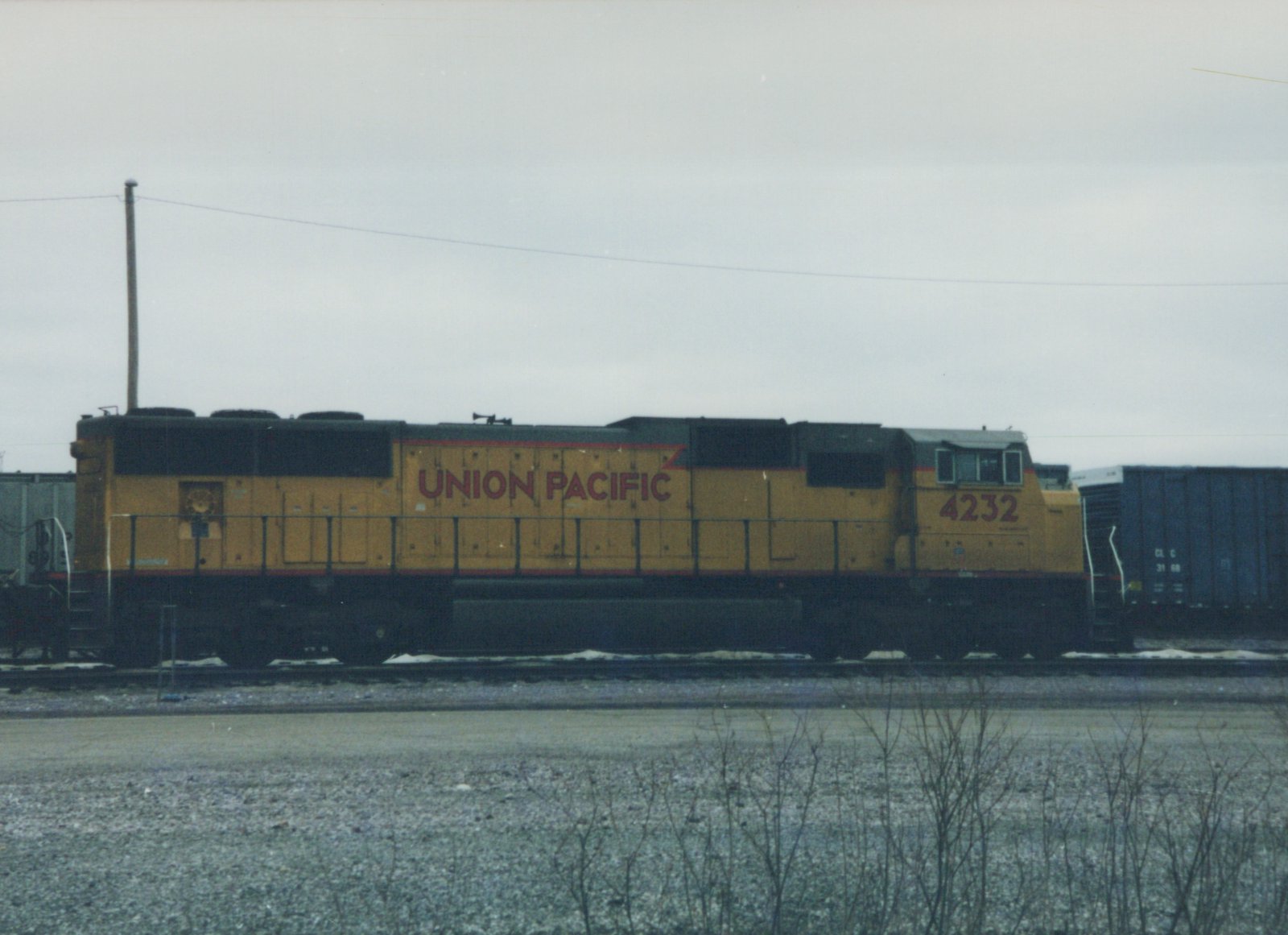 Side view of a yellow Union Pacific diesel locomotive numbered 4232 standing on yard tracks, with a blue hopper car behind it, patches of snow and bare shrubs in the foreground.