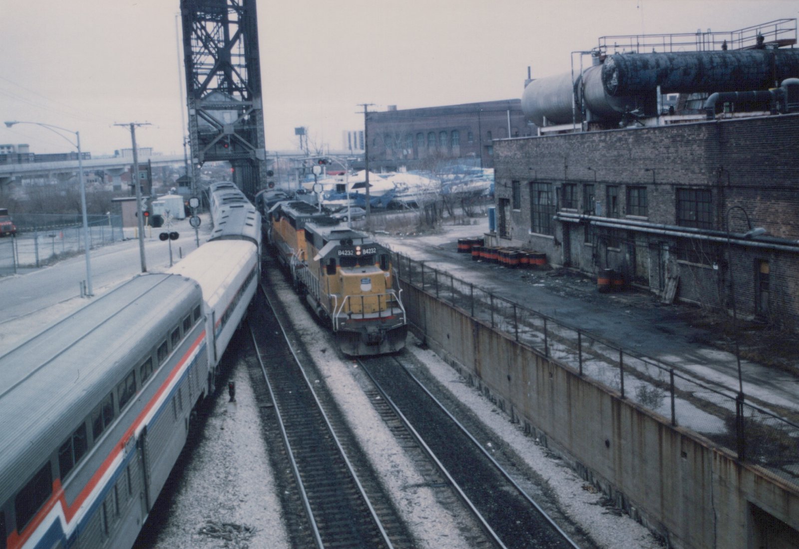 Seen from above, an Amtrak train of stainless and white passenger cars on the left passes a yellow and gray Union Pacific locomotive consist on the right, with a raised steel lift bridge, signals, a brick industrial building and stored boats behind.
