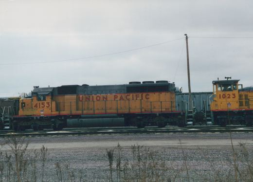 Side view of a yellow Union Pacific road locomotive numbered 4153 and a yellow switcher numbered 1023 on yard tracks under a gray sky, with dry weeds in the foreground.