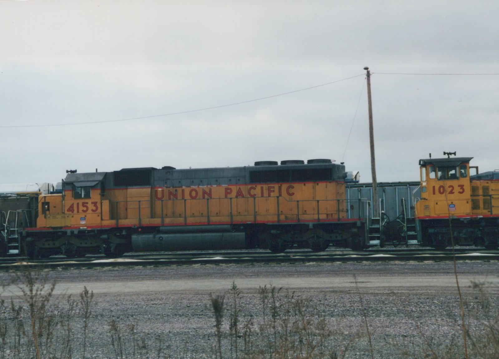 Side view of a yellow Union Pacific road locomotive numbered 4153 and a yellow switcher numbered 1023 on yard tracks under a gray sky, with dry weeds in the foreground.