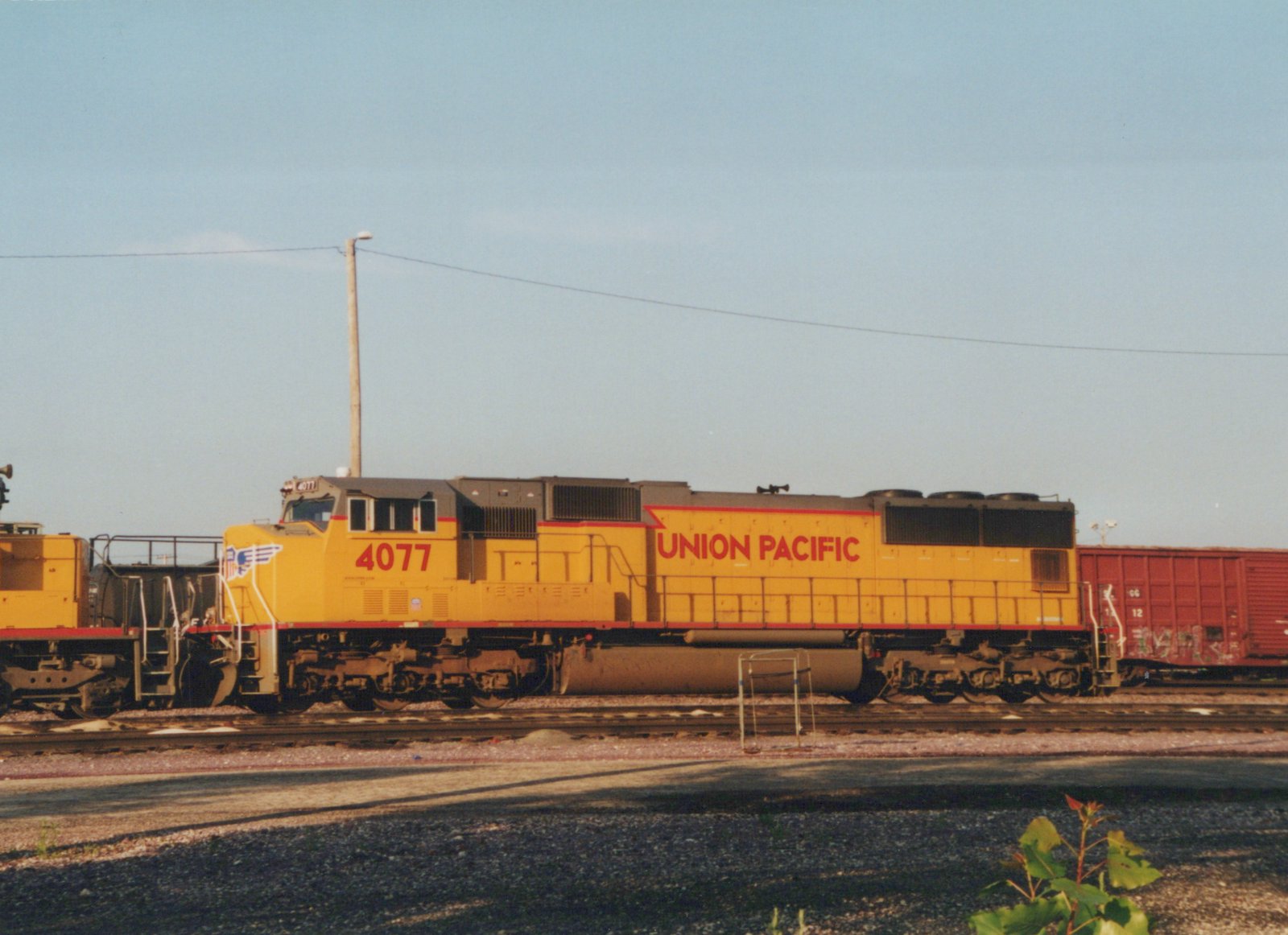 A yellow and gray Union Pacific diesel locomotive numbered 4077, seen from the side in low sunlight on a yard track, with another yellow locomotive coupled at left and a red boxcar with graffiti at right.