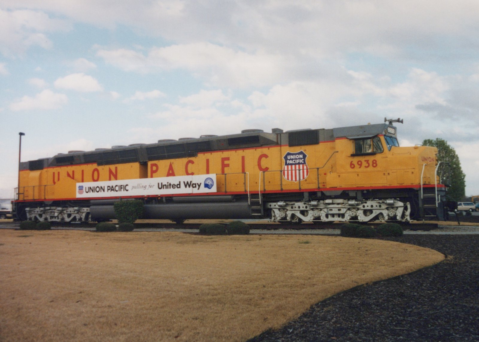 A long yellow Union Pacific diesel locomotive numbered 6938, with red lettering and a shield herald, sits on a short display track behind a brown lawn and trimmed shrubs under a cloudy sky.
