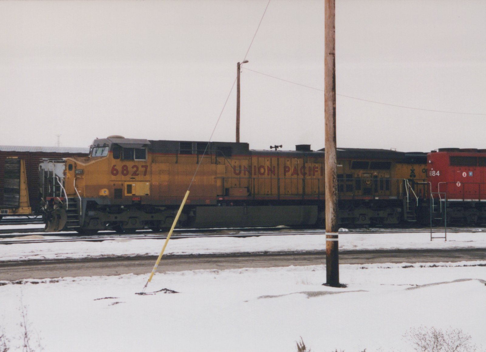 A yellow Union Pacific diesel locomotive numbered 6827, seen from the side on a yard track under a gray sky, with snow on the ground, a wooden utility pole in front and a red locomotive at the right edge.